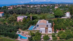 An aerial view of a residential area with a large house featuring a swimming pool, multiple decks, and a view of the sea and mountains in the background. - Alonaki Resort (Preveza)