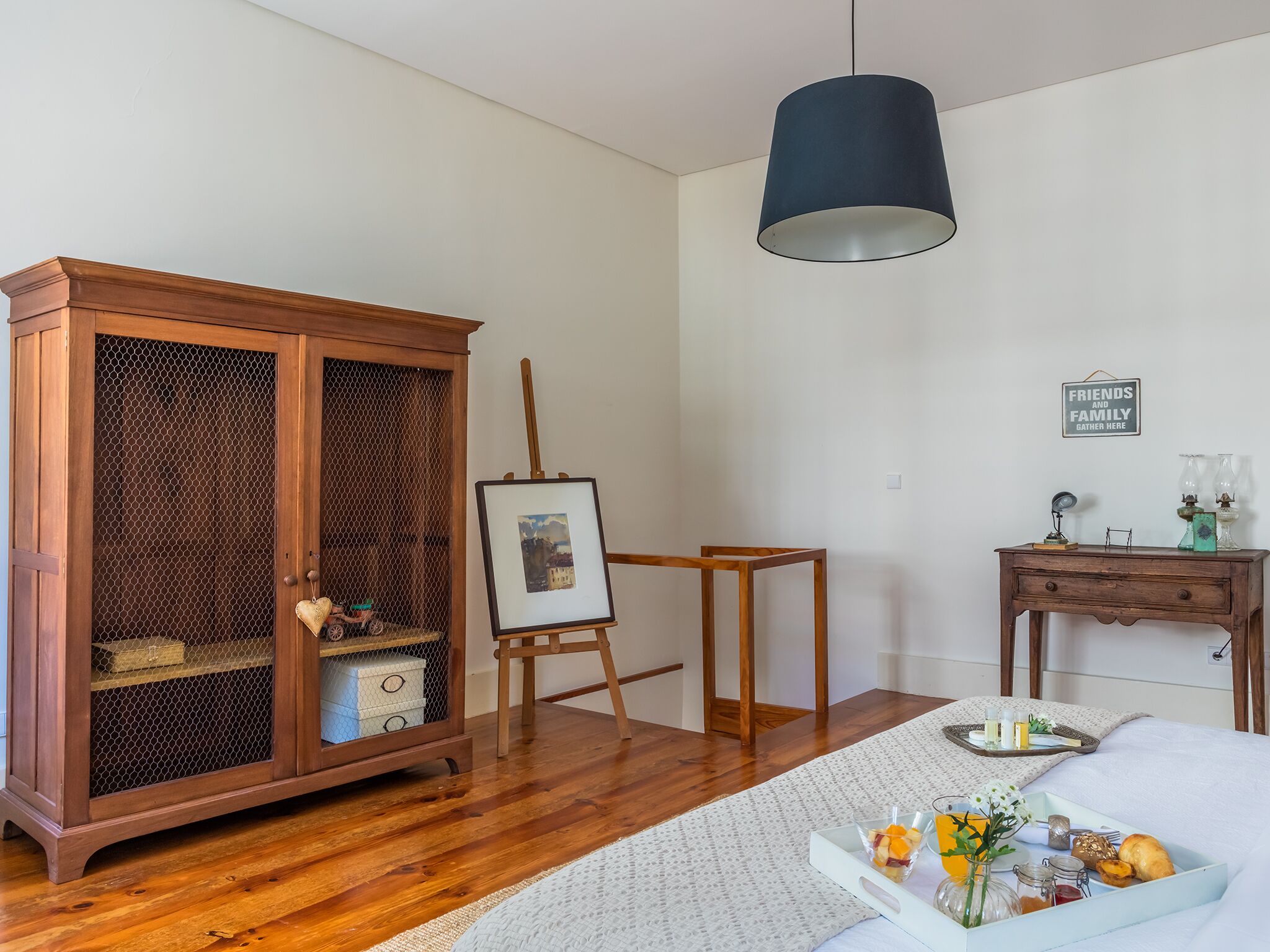 A bedroom with a wooden wardrobe, an easel with a painting, a bedside table with a tray of breakfast items, and a hanging lamp.