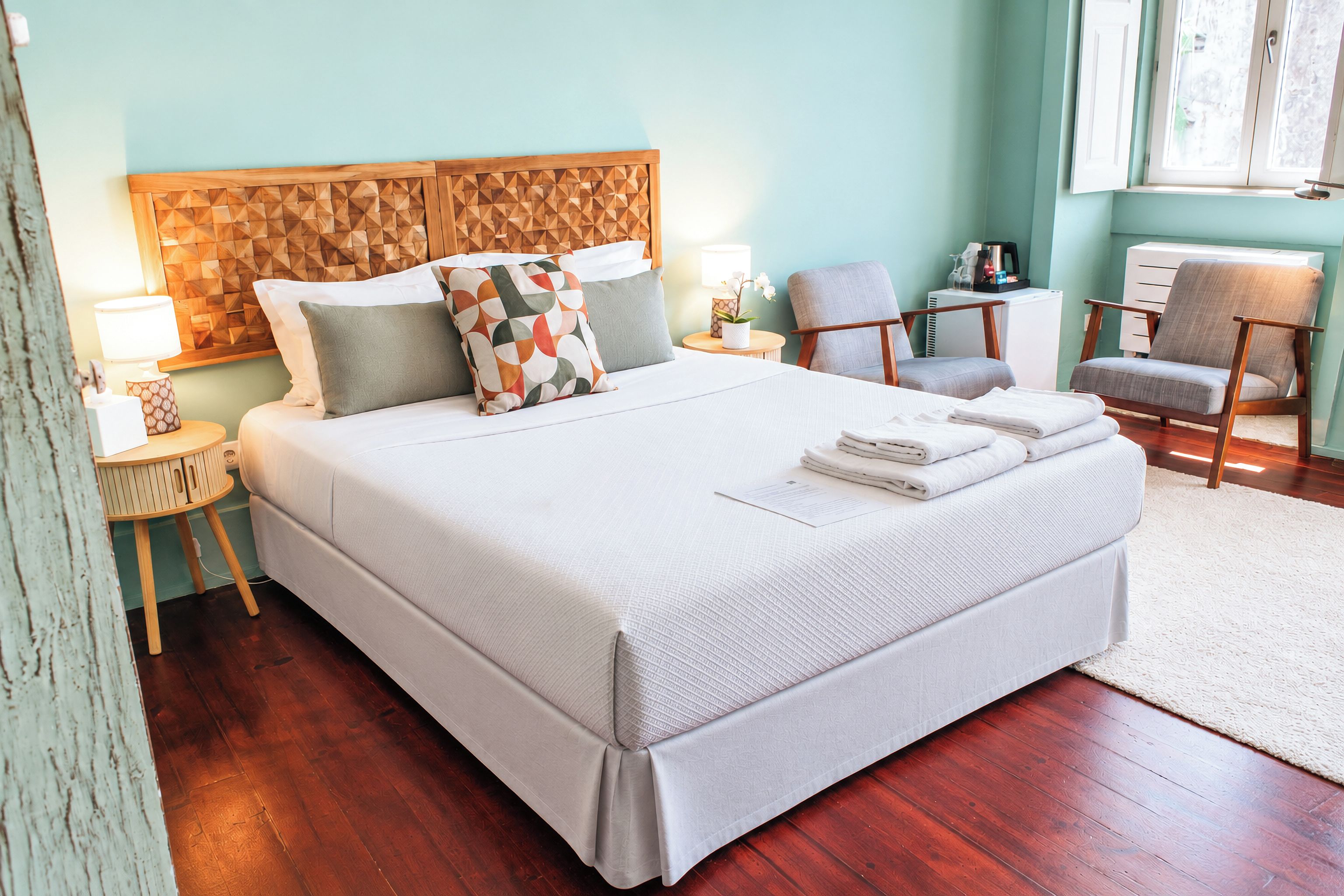 A neatly made bed with pillows, a bedside table with a lamp, a chair, and a radiator in a room with wooden flooring and a light-colored wall.