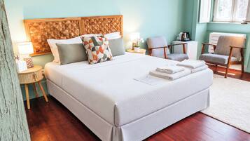A neatly made bed with pillows, a bedside table with a lamp, a chair, and a radiator in a room with wooden flooring and a light-colored wall.