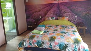 A bedroom with a bed covered in a floral-patterned bedspread, a wall mural of purple flowers, and a small wooden nightstand with a vase. - Vacation home for 10 people 1h15 from Paris (Venon)