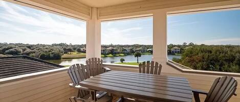 A covered patio with a wooden table and chairs, large windows offering a view of a lake and surrounding greenery.