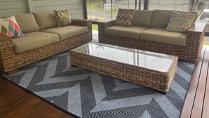 Een rieten zithoek met kussens, een salontafel met glazen blad en een kamerkleed op een houten terras.
