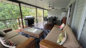 Een overdekt terras buiten met een eettafel, stoelen, een barbecue en rieten meubilair.
