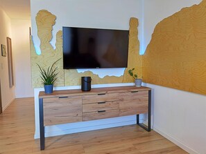 A modern living room with a flat-screen TV mounted on a textured wall, a wooden console table with drawers, and two potted plants.