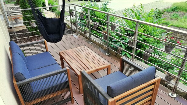 Um deck de madeira com um sofá e uma mesa, cercado por vasos de plantas e um corrimão de metal.