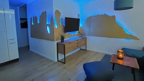 Uma sala de estar moderna com um móvel de TV de madeira, uma TV de tela plana, uma luminária fixada na parede e uma mesinha com uma vela.