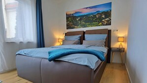 Uma cama com cabeceira, duas luminárias de apoio, uma janela com cortinas e um quadro de paisagem na parede.