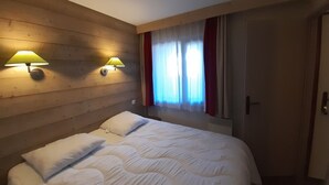 Quarto de hotel com cabeceira de madeira, duas luminárias fixadas na parede, cama com roupa de cama branca, janela com cortinas leves e uma porta.