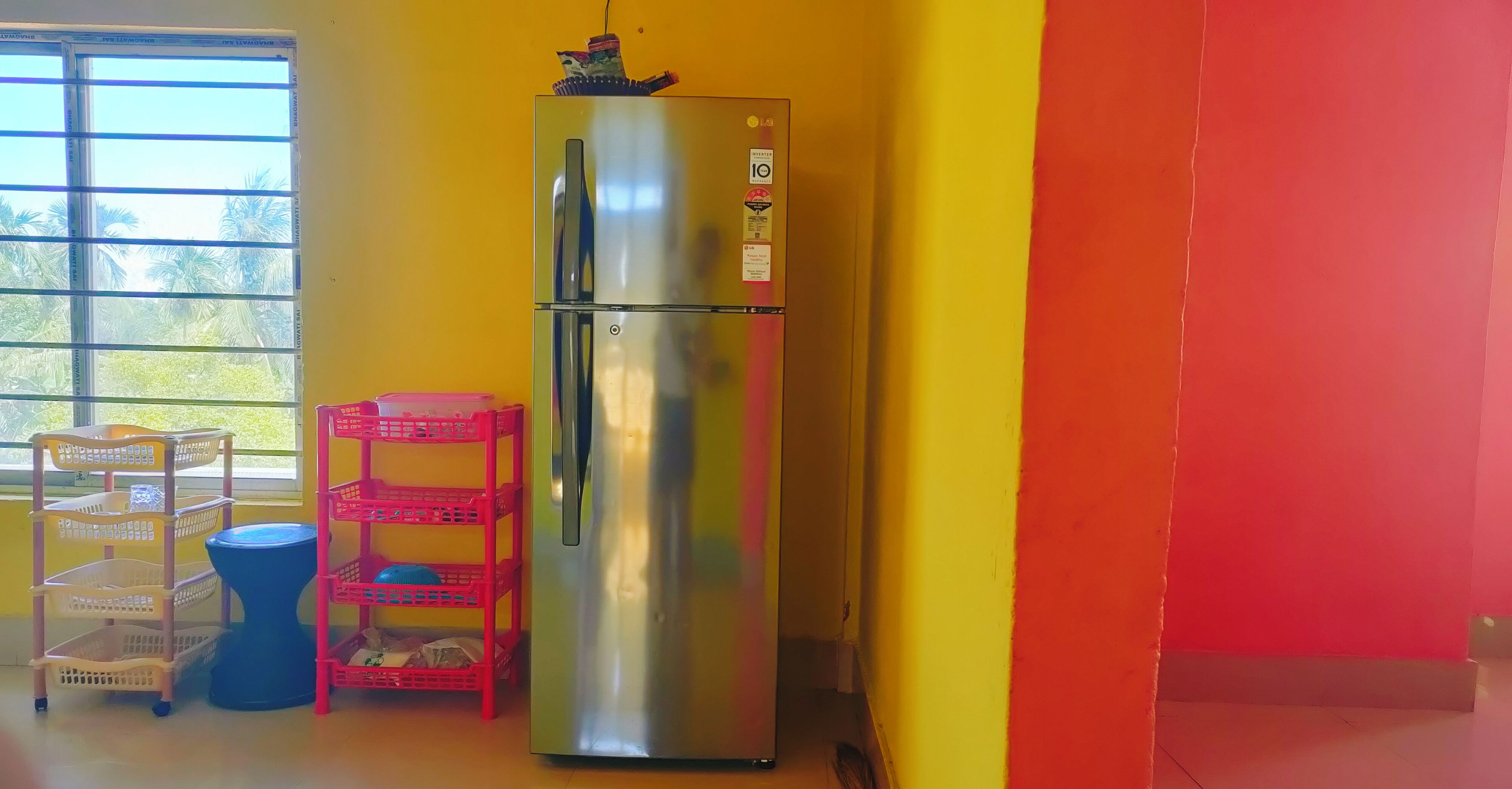 A stainless steel refrigerator in a room with yellow and orange walls, a window with blinds, and a blue trash bin.