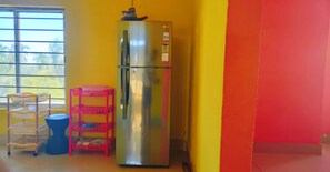 A stainless steel refrigerator in a room with yellow and orange walls, a window with blinds, and a blue trash bin. - Couple friendly space. (Tezpur)