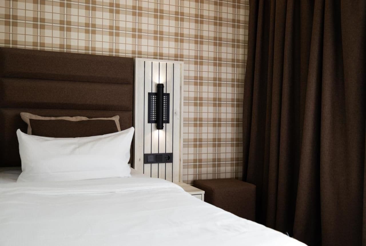 A hotel room with a bed, a nightstand, a wall-mounted control panel, and a curtain.