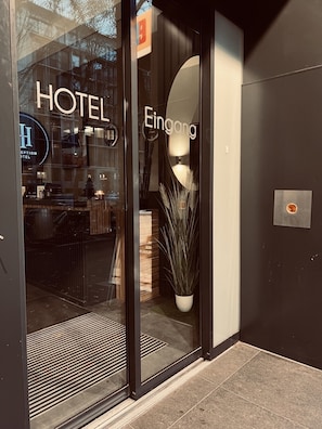 A hotel entrance with a reception desk, a potted plant, and a sign reading "HOTEL Eingang".