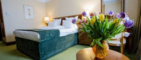 A hotel room with a bed, a wooden nightstand, a vase with flowers, and a mirror on the wall.