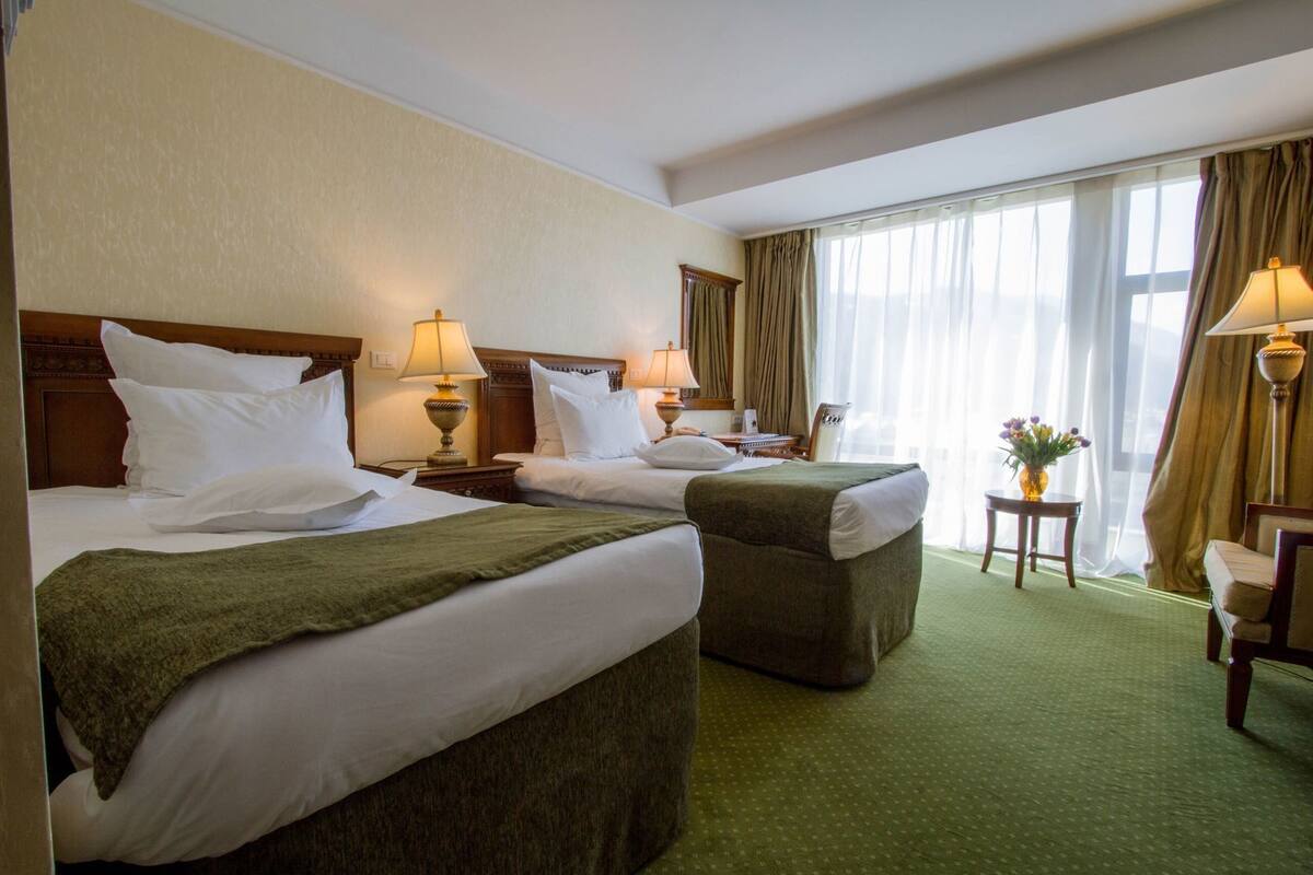 A hotel room with two beds, a green carpet, a chair, a table with a vase, and a window with curtains.