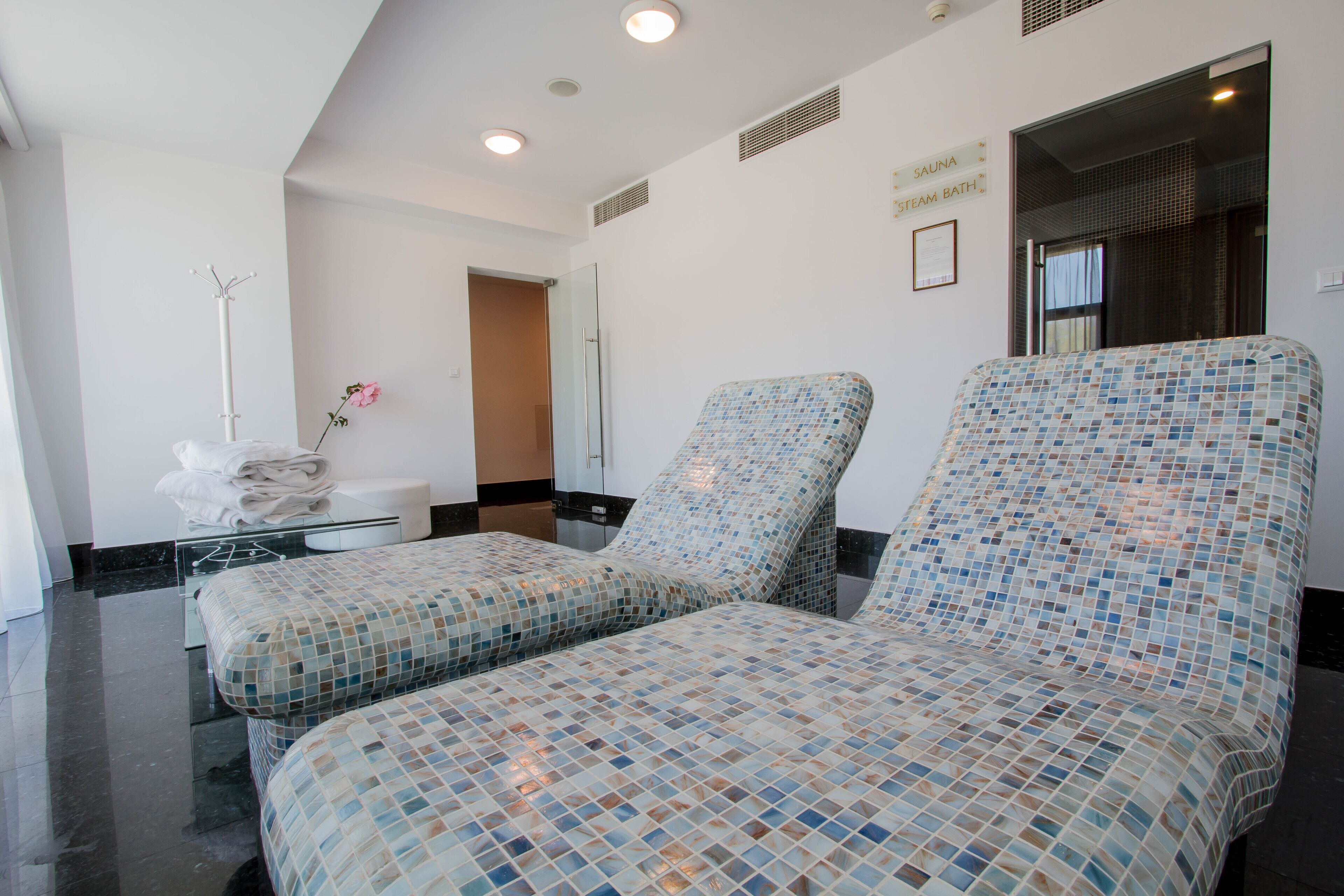 A spa room with two mosaic-tiled massage chairs, a glass table with towels, and a sauna sign on the wall.