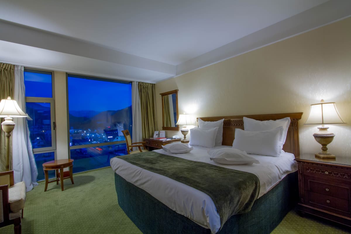 A hotel room with a large bed, two bedside lamps, a wooden nightstand, a small table, and a view of a cityscape through a large window.