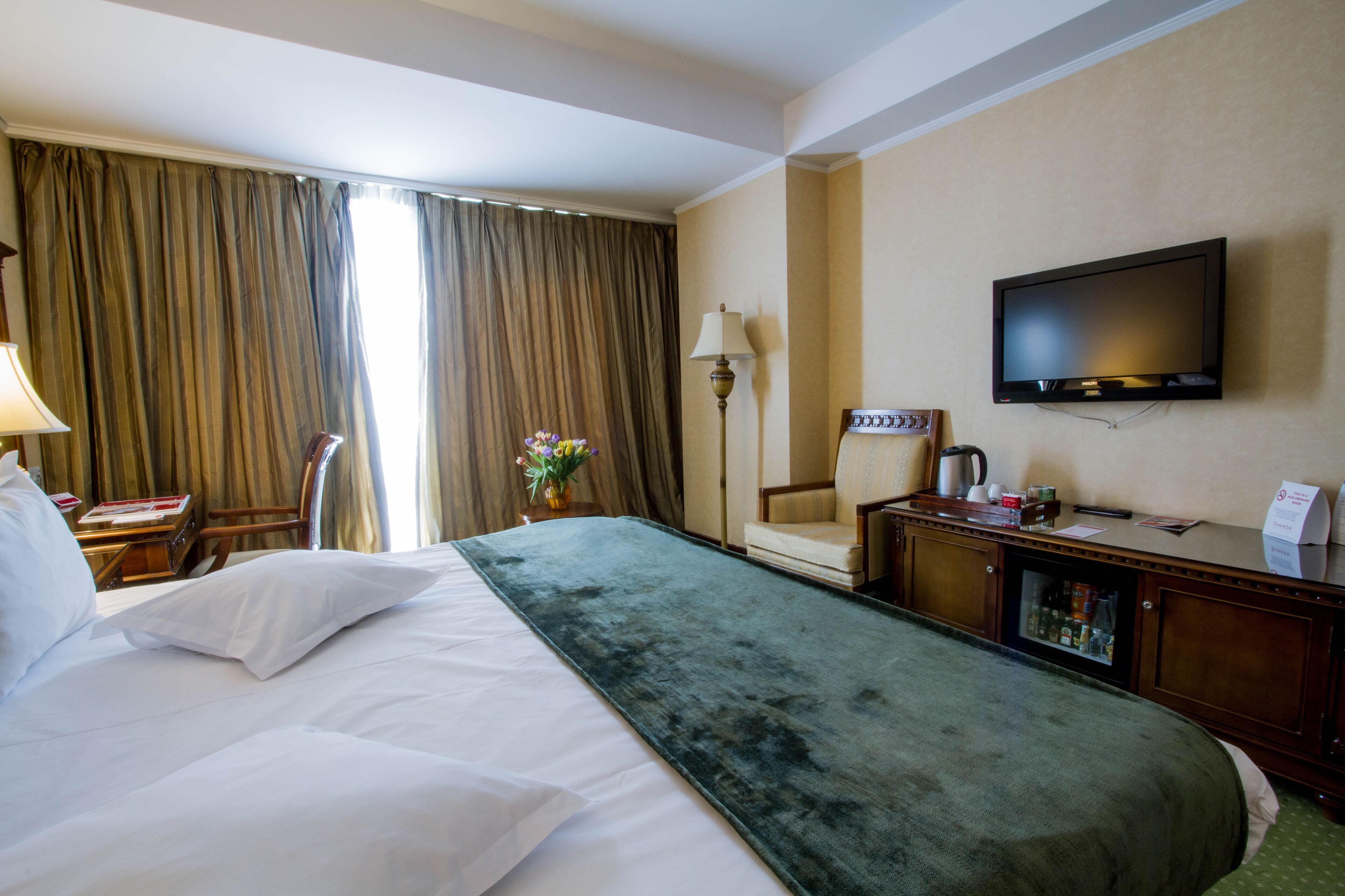 A hotel room with a large bed, a desk with a lamp, a television, and a minibar.