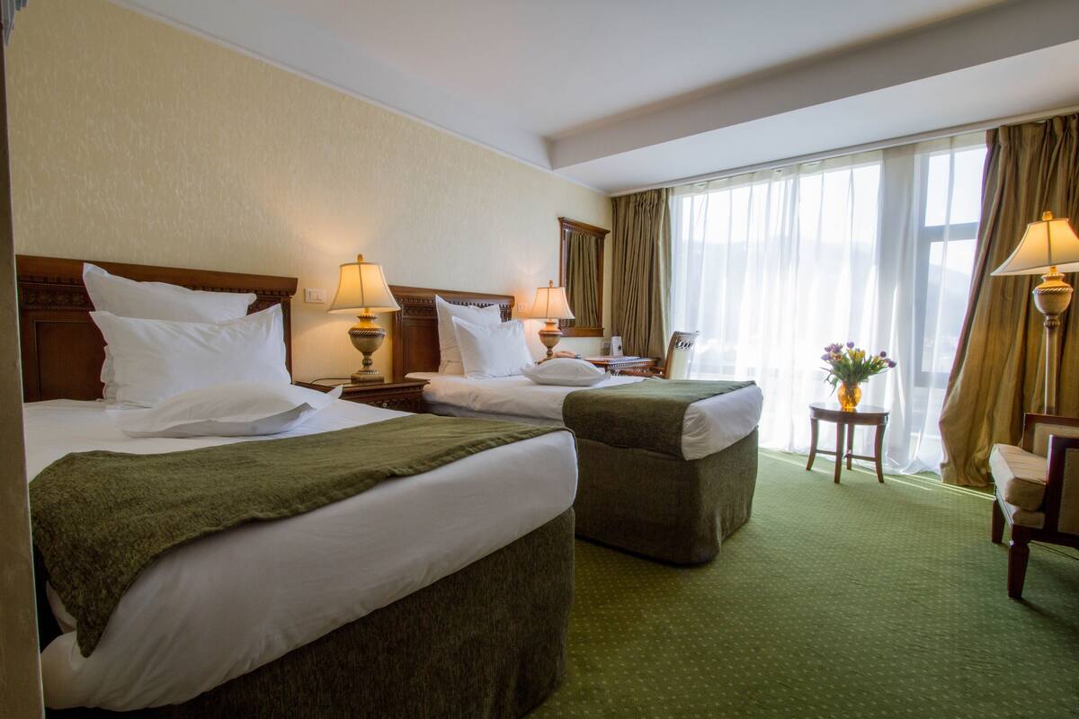 A hotel room with two beds, a green carpet, a small table with a vase, and a chair.