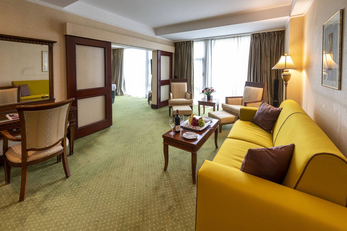 A hotel room with a yellow sofa, a wooden chair, a small table with a tray of fruit and a bottle, and a lamp.