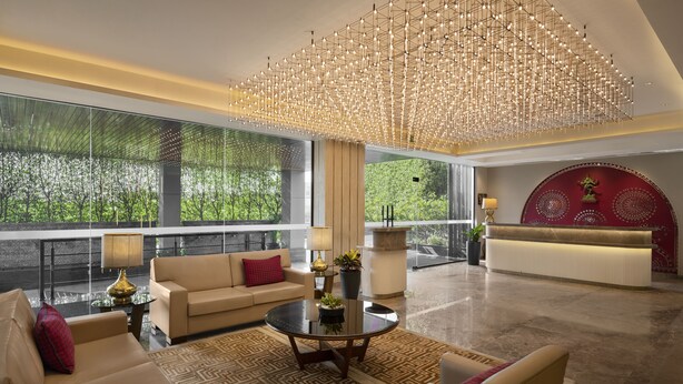 A spacious lobby with a modern chandelier, a reception desk, and a large window offering a view of greenery. - The Neemaya Hotel (Gandhidham)