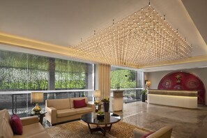 A spacious lobby with a modern chandelier, a reception desk, and a large window offering a view of greenery. - The Neemaya Hotel (Gandhidham)