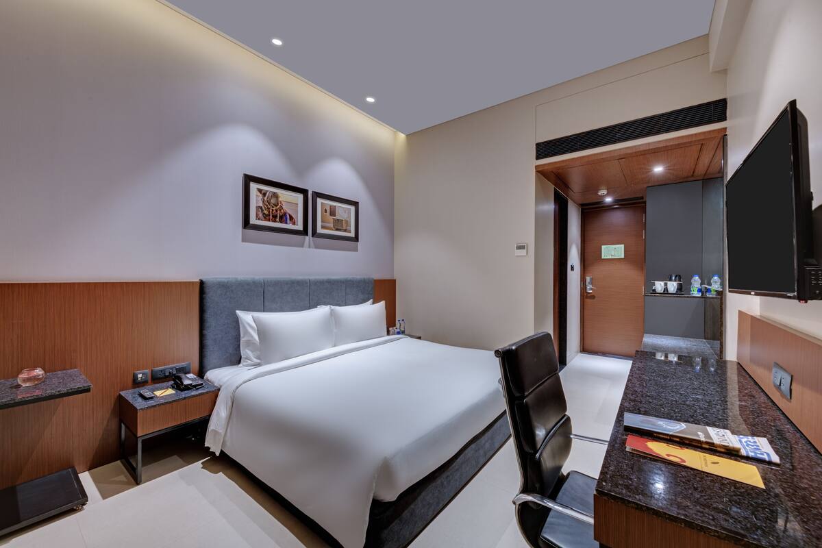 A modern hotel room with a neatly made bed, a desk with a chair, a flat-screen TV, and a small kitchenette.