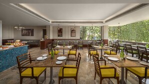 A restaurant with multiple tables and chairs, a buffet area, and large windows offering a view of greenery. - The Neemaya Hotel (Gandhidham)