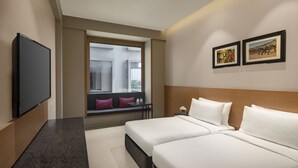A hotel room with two beds, a TV, a window with a view of buildings, and two framed pictures on the wall. - The Neemaya Hotel (Gandhidham)