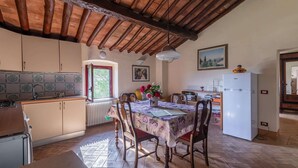 A kitchen with wooden cabinets, a dining table with chairs, a refrigerator, and a window with a view of greenery. - Torriano 2 (San Casciano Val di Pesa)