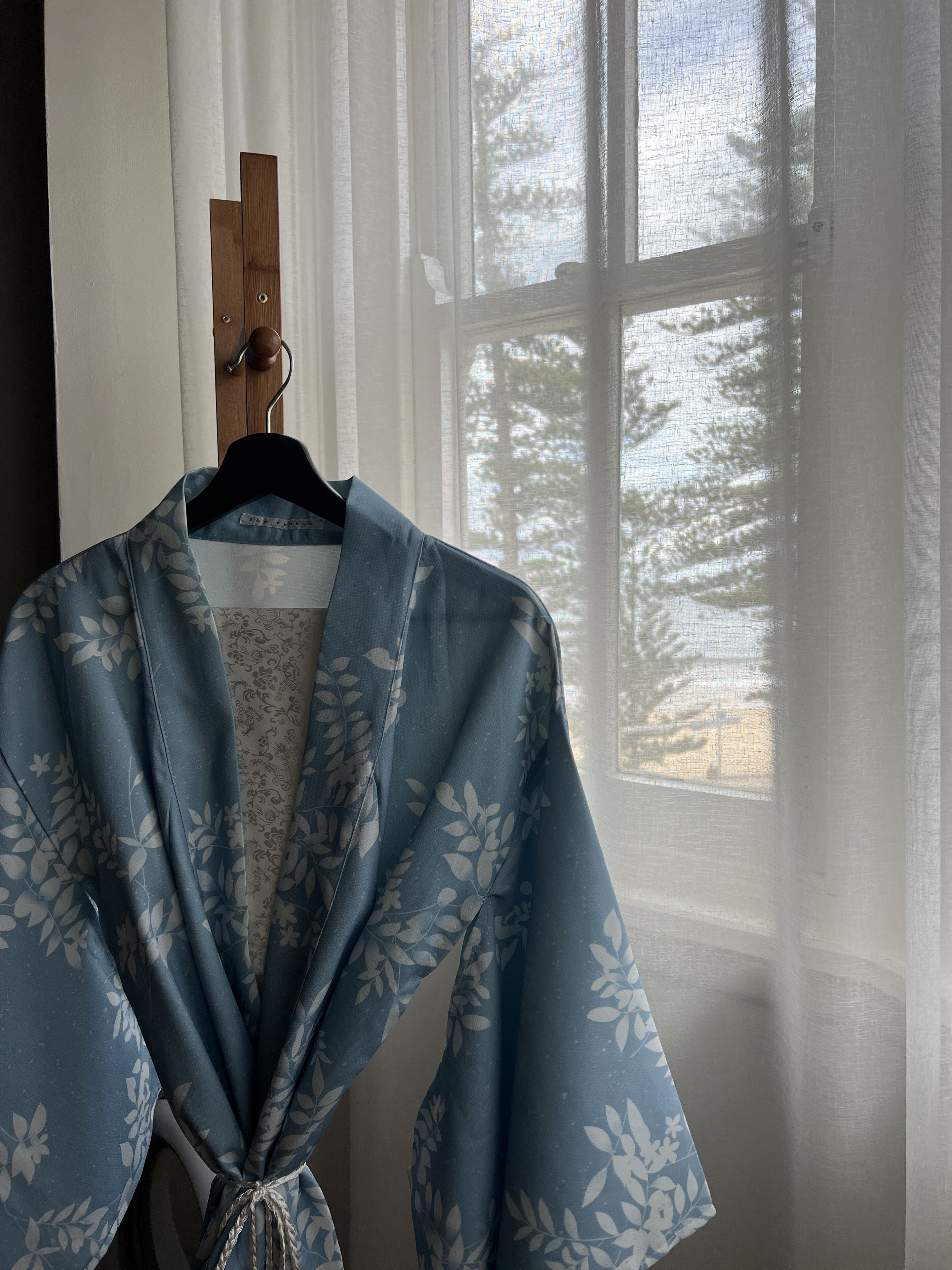 Un kimono bleu orné d’un motif floral est suspendu à un cintre en bois près d’une fenêtre dont les rideaux sont en voile léger.