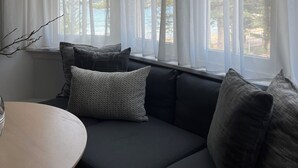 A modern living room with a dark gray sofa, white curtains, and a view of trees through large windows. - The Ryokan - Zen (Manly)