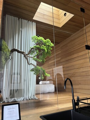Un intérieur moderne avec un plafond en bois, une lucarne, un bonsaï en pot, un rideau blanc, un robinet noir et un panneau encadré posé sur le sol.