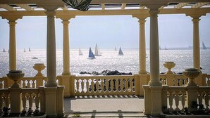 Vista al mar desde un balcón cubierto con columnas y barandillas, con un mar tranquilo y veleros.
