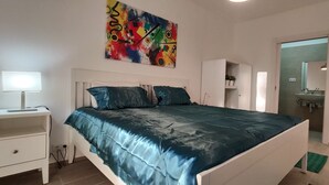 Un dormitorio con cama, mesita de noche, lámpara, armario y baño visible a través de una puerta abierta.
