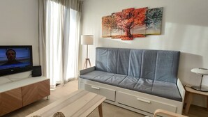 Una sala de estar con un sofá gris, un televisor sobre un mueble de madera, un cuadro con hojas otoñales y una lámpara en una mesita.
