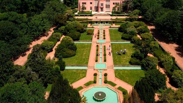 Vista aérea de un jardín formal con una fuente central, senderos simétricos y un edificio grande al fondo.