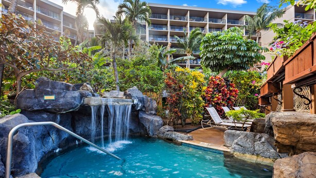 Zona de piscina con cascada, rodeada de plantas tropicales y un edificio de hotel de varios pisos al fondo.
