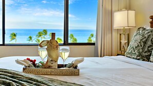 Una cama bien tendida con una bandeja de copas de vino, una botella y frutas, con vistas a un paisaje marino a través de amplios ventanales.