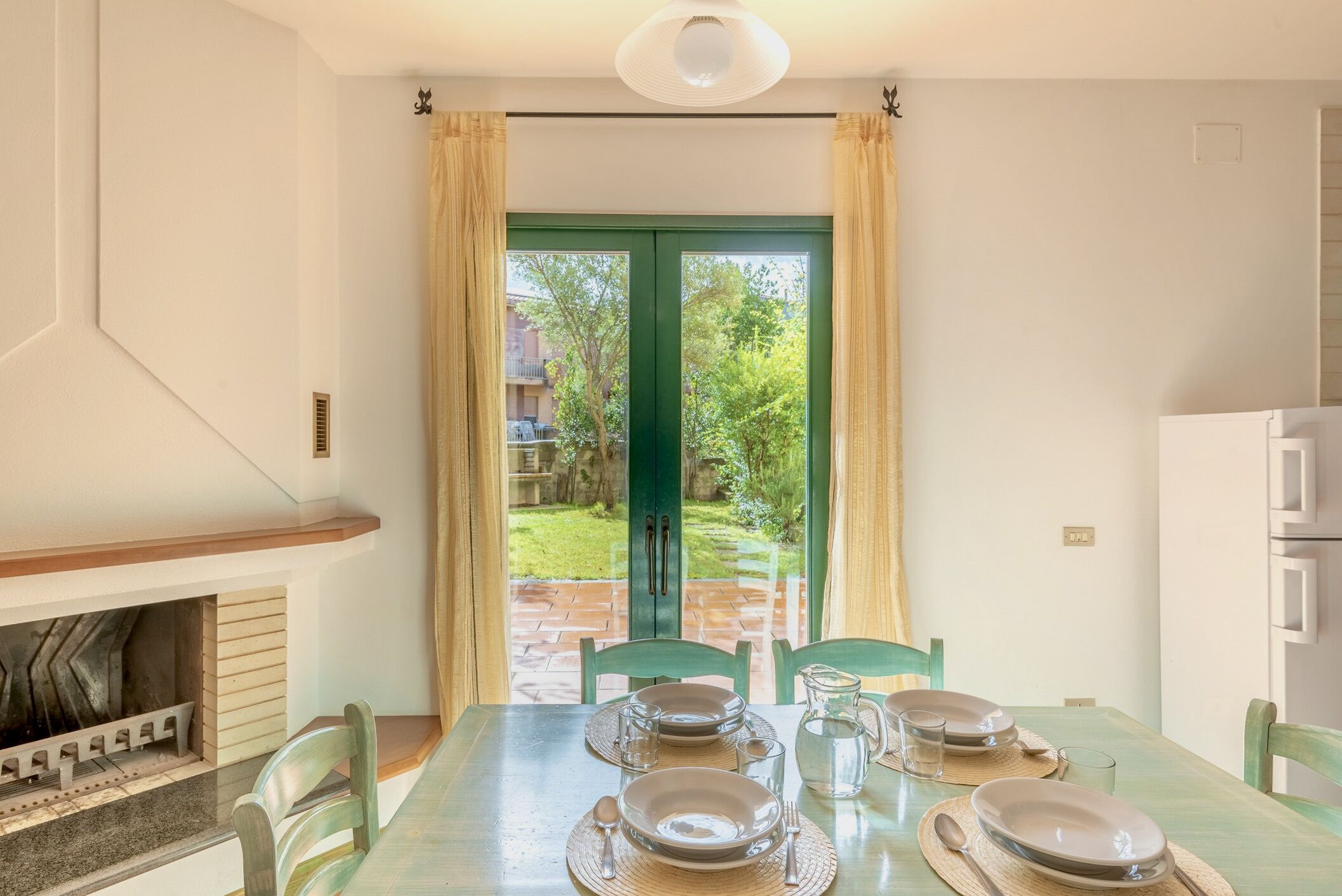 A dining area with a table set for four, a fireplace, and a view of a garden through a green door.