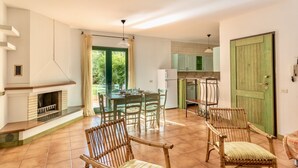 A spacious living area with a fireplace, dining table, and chairs. There is a kitchen area with cabinets and a green door. - I Mirti Bianchi Residence con piscina (Santa Teresa di Gallura)