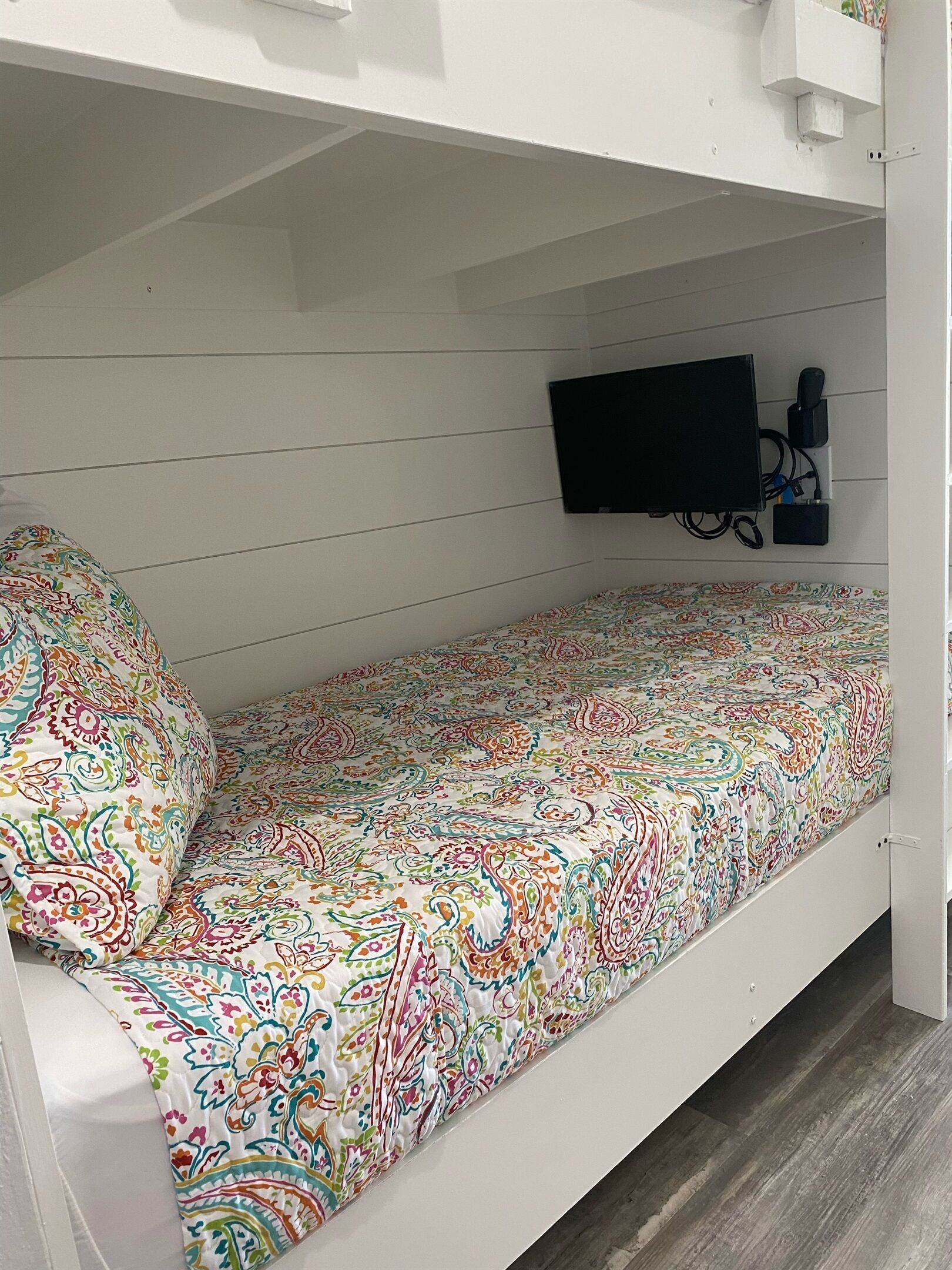 Una cama litera con una colcha colorida, un televisor montado y un enchufe.
