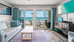 En stue med sofa, sofabord og fjernsyn, med udsigt over en strand og en balkon.