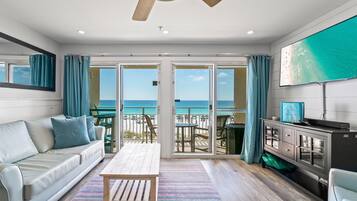 A living room with a sofa, coffee table, and TV, overlooking a beach with a balcony.
