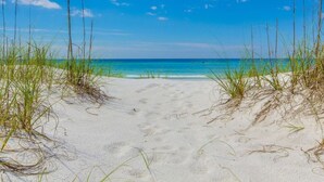 En strand med klart, blåt vand og hvidt sand, omgivet af højt græs.