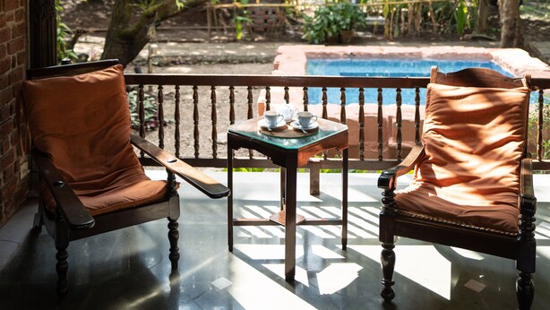 A covered patio with two leather chairs and a table, overlooking a swimming pool and lush greenery. - Amã Stays & Trails Asanji Wadi (Alibag)