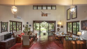 A spacious living area with a dining table, chairs, a sofa, and a coffee table. There is a large window with a view of greenery outside. - Amã Stays & Trails Asanji Wadi (Alibag)