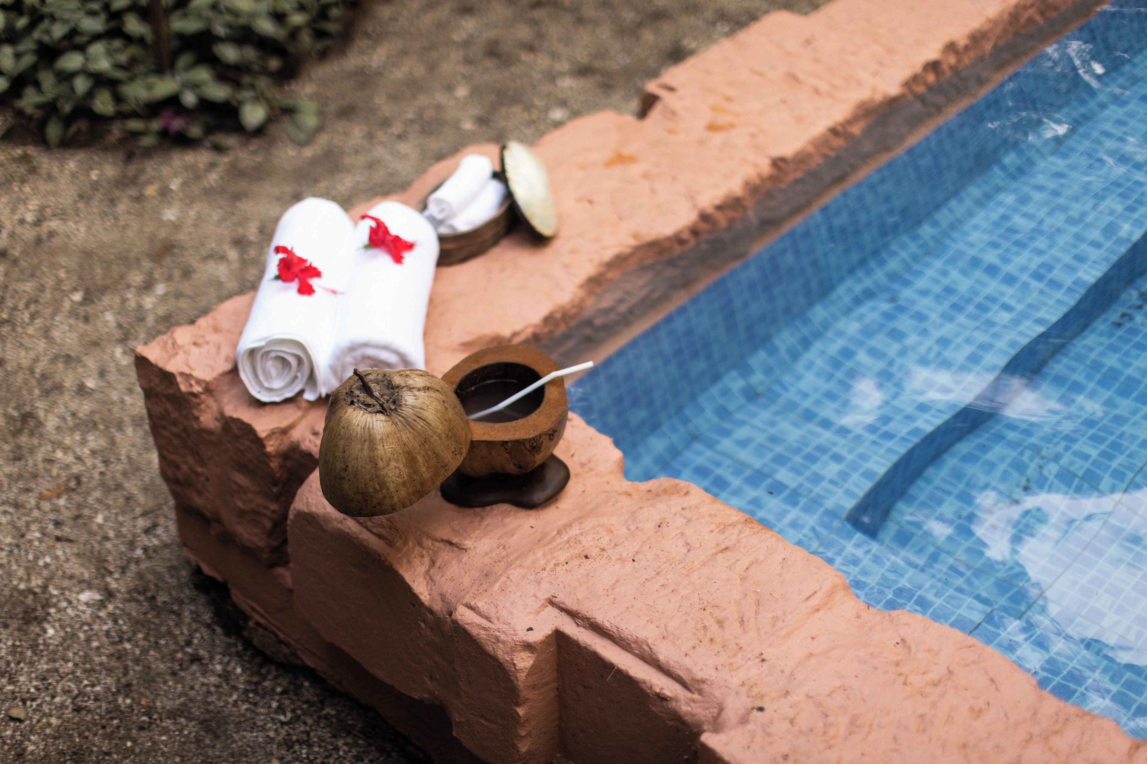 Un espace aménagé au bord de la piscine, avec une bordure en briques, une serviette ornée d’une fleur rouge et une noix de coco.