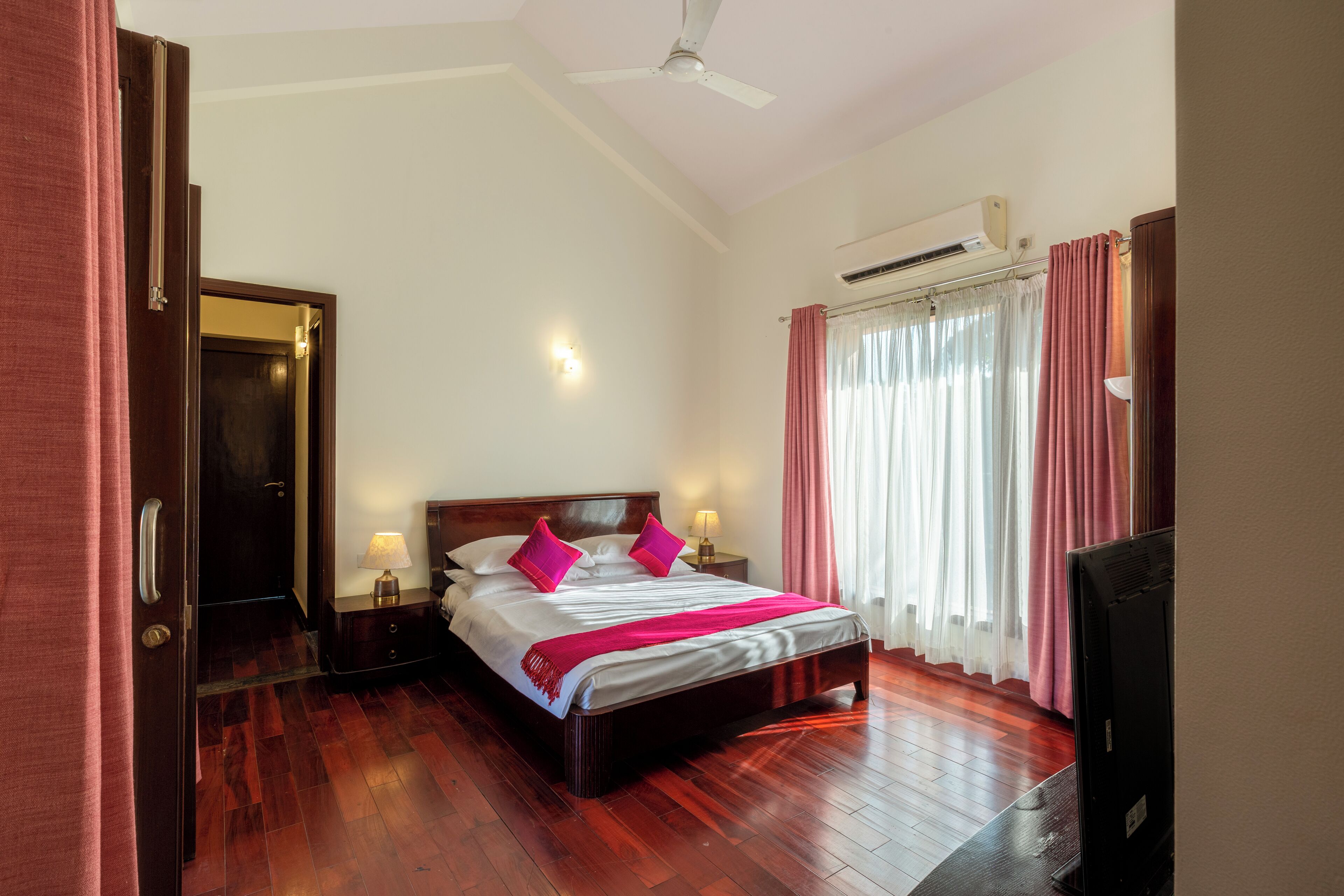 Habitación de hotel con piso de madera, cama con ropa blanca y almohadas rosas, mesita de noche con lámpara, televisor y aire acondicionado.