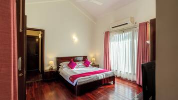 Habitación de hotel con piso de madera, cama con ropa blanca y almohadas rosas, mesita de noche con lámpara, televisor y aire acondicionado.